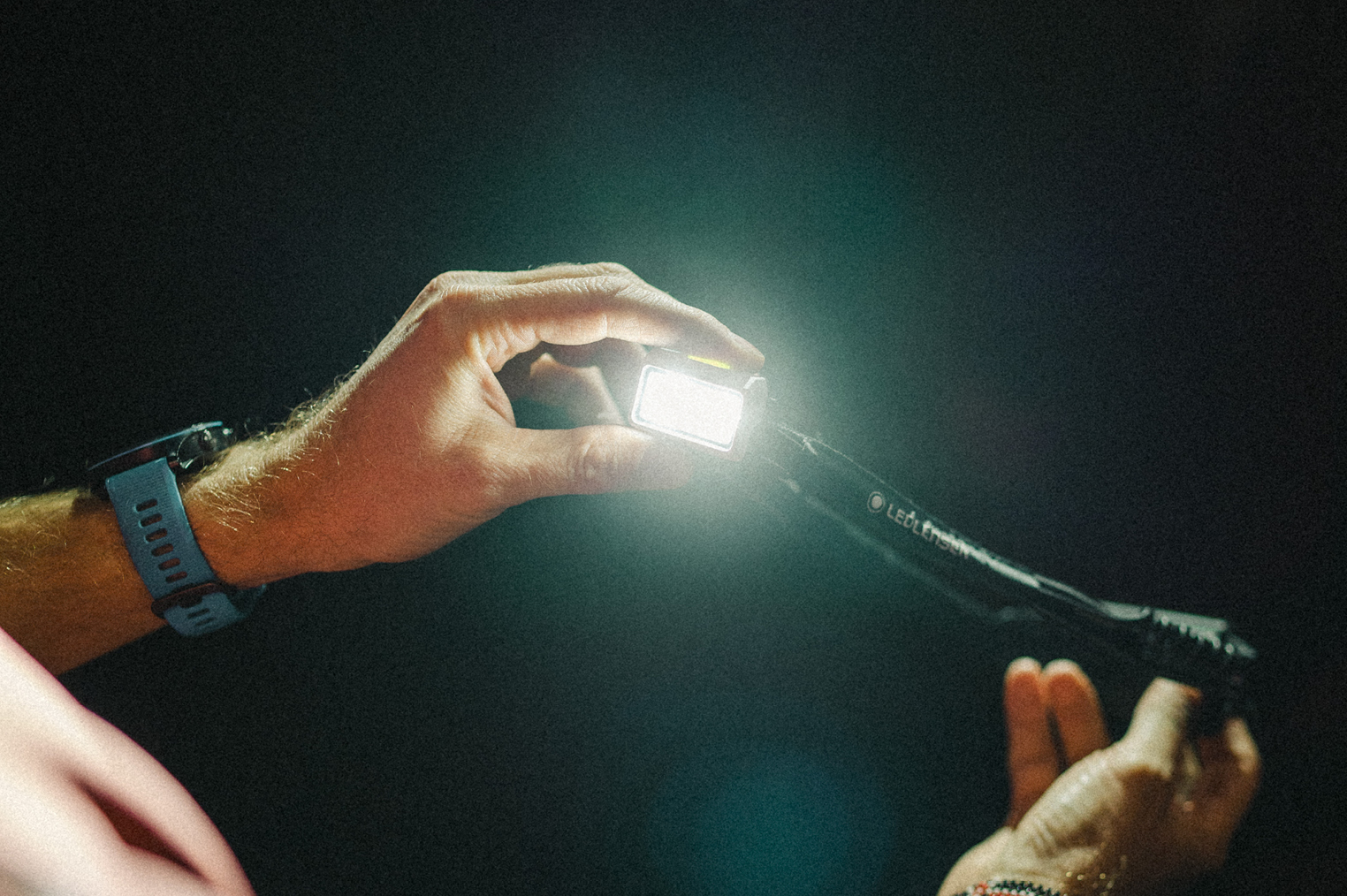 มือถือไฟฉายคาดหัว Ledlenser เปิดไฟสว่างในความมืด (Hands holding a lit Ledlenser headlamp in the dark)