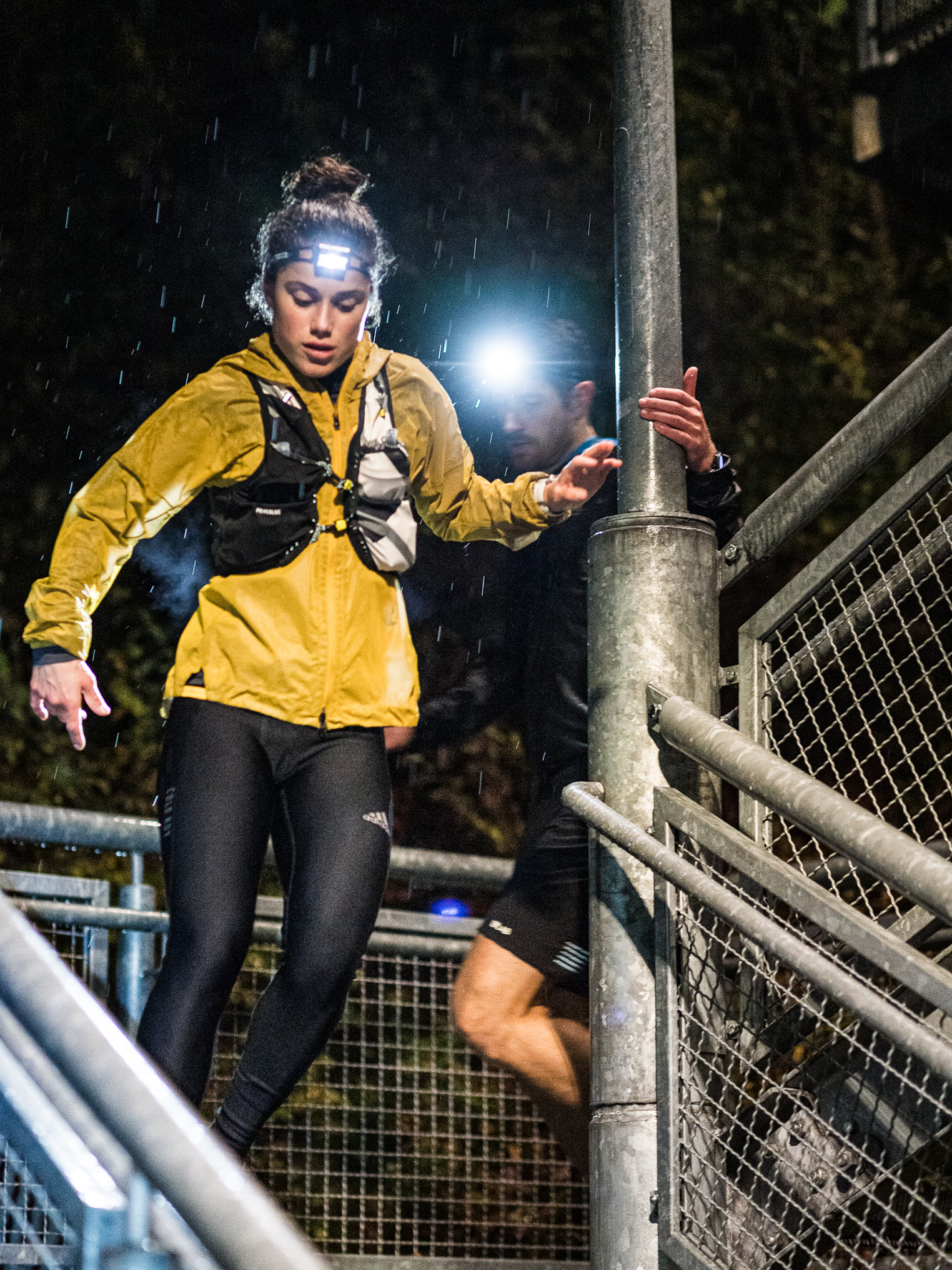 นักวิ่งหญิงสวมไฟฉายคาดหัว Ledlenser วิ่งลงบันไดกลางสายฝนยามกลางคืน (Female runner wearing a Ledlenser headlamp descending stairs in the rain at night)