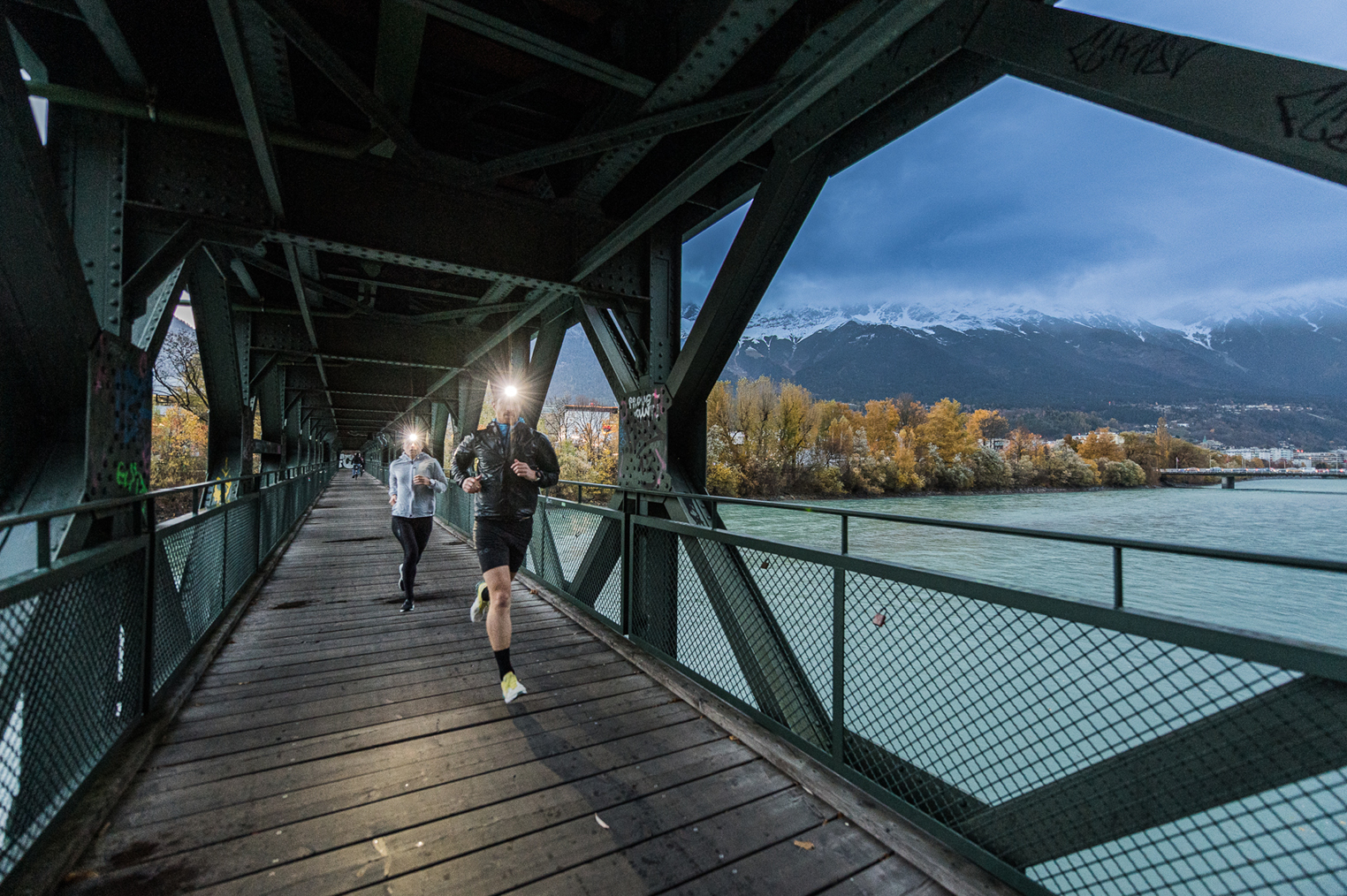 นักวิ่งสองคนสวมไฟฉายคาดหัว Ledlenser วิ่งข้ามสะพานยามพลบค่ำ (Two runners with Ledlenser headlamps crossing a bridge at dusk)