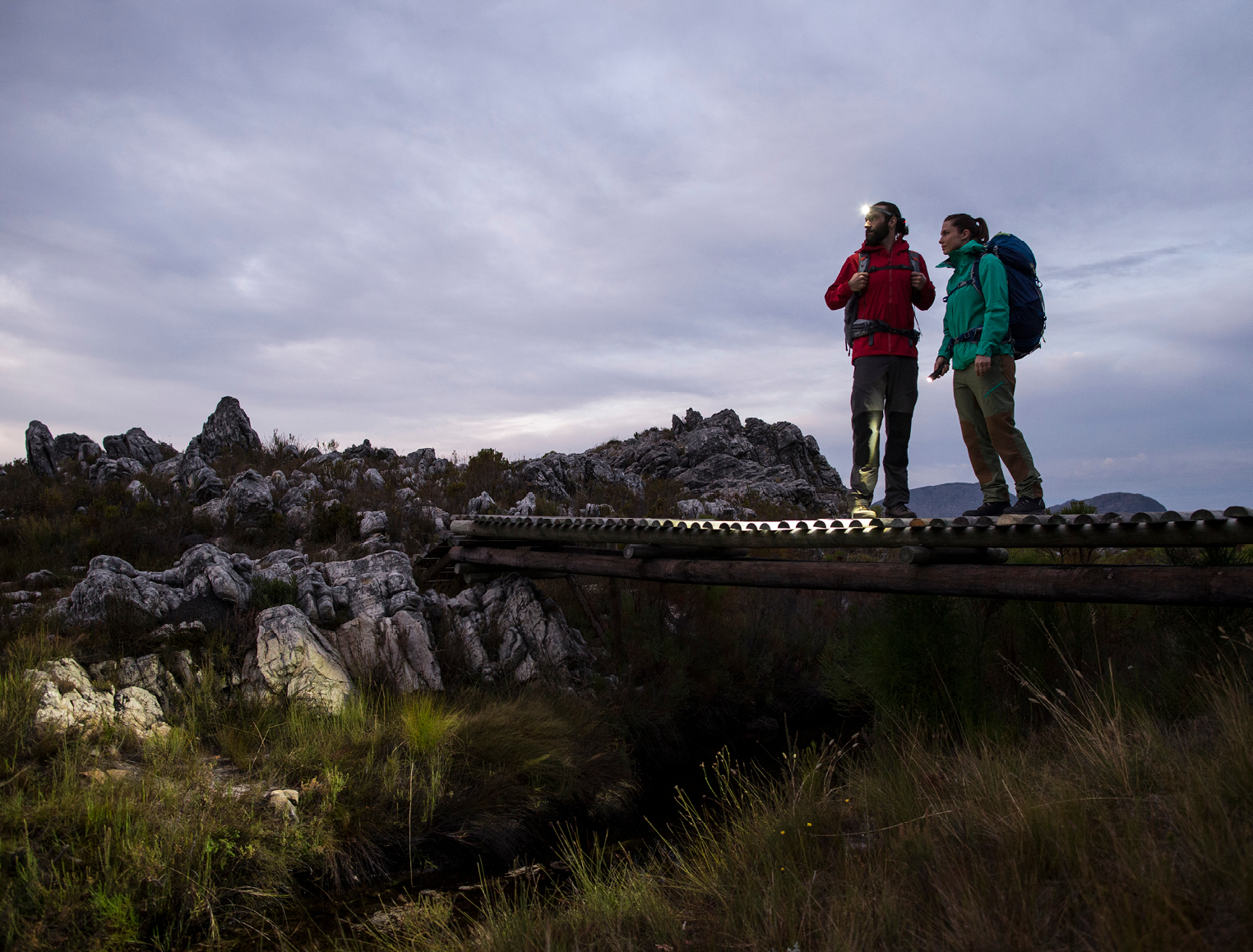 นักเดินป่าชายสวมไฟฉายคาดหัว Ledlenser เปิดไฟยืนบนสะพานไม้ยามพลบค่ำ (Male hiker wearing a lit Ledlenser headlamp on a wooden bridge at dusk)
